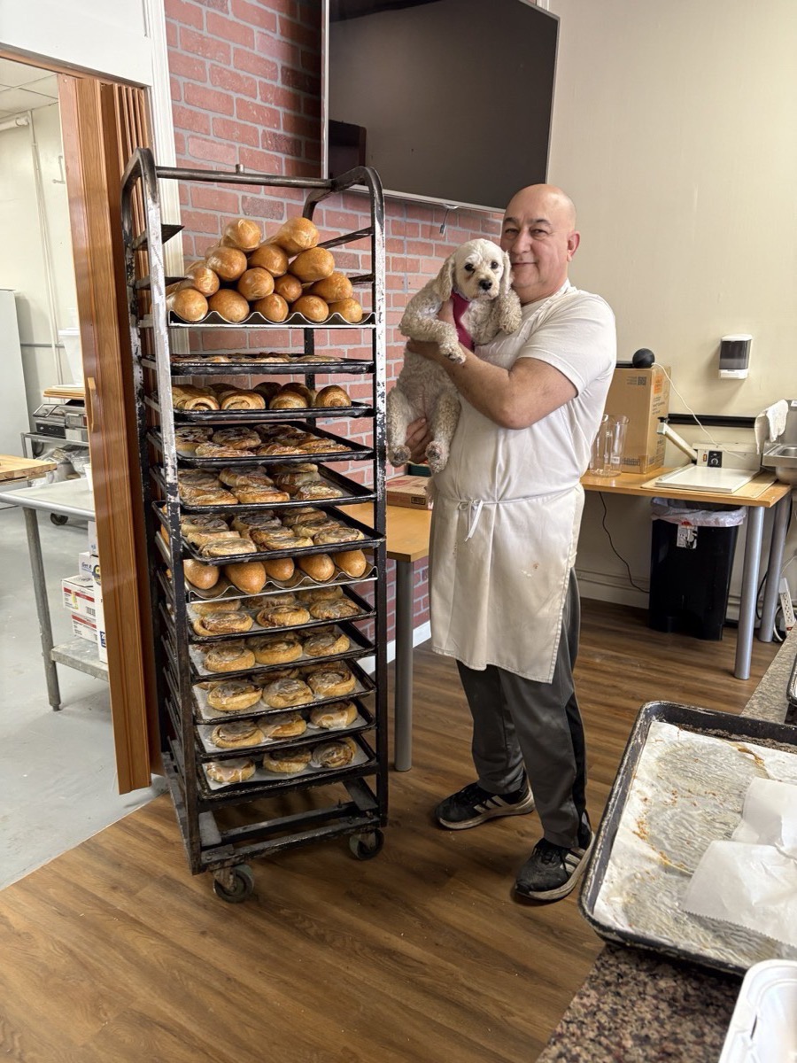 Master Baker Max Malek with his dog beside fresh morning bread and pastry racks at Just Baked Bakery